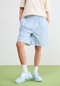 Pantaloni corti a righe blu e bianche con cintura elasticizzata, abbinati a scarpe slip-on di colore blu chiaro e calzini bianchi alla caviglia. Sfondo verde.
