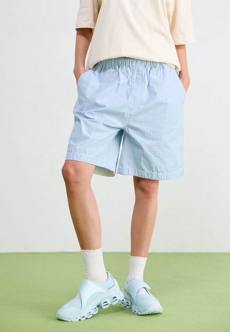 Pantaloni corti a righe blu e bianche con cintura elasticizzata, abbinati a scarpe slip-on di colore blu chiaro e calzini bianchi alla caviglia. Sfondo verde.