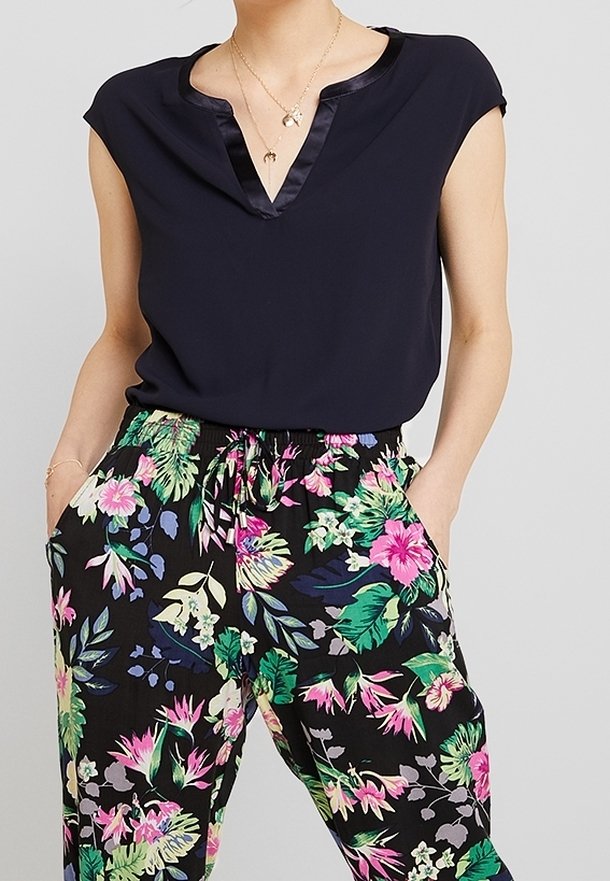Personne portant un haut noir à manches courtes avec une bordure en satin et un pantalon noir taille haute avec un imprimé floral tropical coloré. Mains dans les poches.