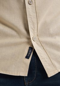 Beige linnen shirt met een knoopsluiting, voorzien van twee knopen. Etiket met "PETROL IND." zichtbaar aan de onderkant, gecombineerd met donkerdenim.