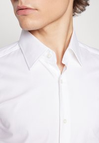 Camicia bianca con bottoni, caratterizzata da un motivo testurizzato, con colletto classico e bottoni a contrasto, parzialmente infilata nella zona del collo.