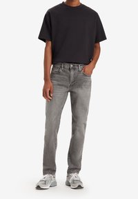 Homme portant un T-shirt noir à manches courtes, un jean gris délavé et des baskets grises et blanches, debout avec une main dans la poche.
