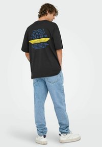 Sort oversized t-shirt med et blåt og gult grafisk print på ryggen, som viser turnédatoer. Sammen med lyseblå denim jeans og hvide sneakers.