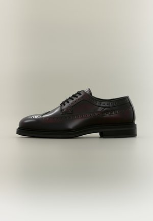 Chaussure habillée brogue en cuir noir avec design wingtip, détails perforés et semelle noire sur fond neutre.