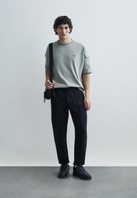 Grå t-shirt med korte ærmer og et diskret logo, kombineret med sorte skræddersyede bukser og sorte sneakers, står på en fast overflade.