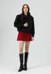 Zwart cropped jasje met opgepofte mouwen en knoopsluiting, gecombineerd met een rode mini jurk en zwarte kniehoge laarzen met hak.