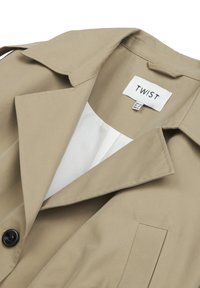 Beiger Trenchcoat mit klassischem Kragen, schwarzem Knopf und weißem Innenfutter. Etikett trägt die Aufschrift "TWIST". Merkmale sind ein minimalistisches Design und eine taillierte Form.