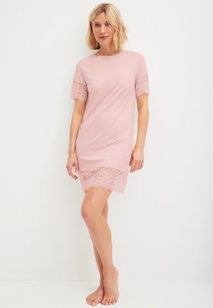 Blonde Frau steht barfuß da und trägt ein kurzes, weiches rosa Kleid mit Spitzensäumen an Ärmeln und Saum vor einem einfarbigen weißen Hintergrund.