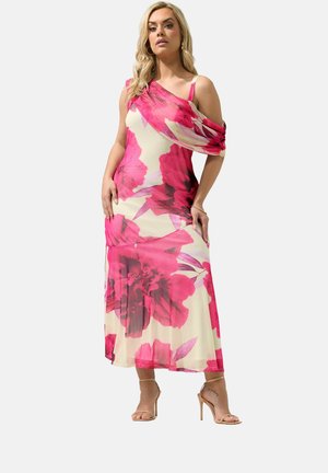 Kvinde iført en ærmeløs creme maxi-kjole med stort pink blomsterprint og asymmetrisk draperet skulder, stående i nøgne højhælede sko.