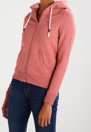 Femme portant un sweat à capuche rose zippé avec des cordons blancs et des poches avant, associé à un jean bleu foncé.