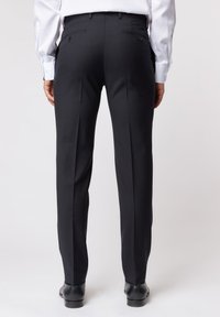 Czarne spodni z dostosowanym krojem o gładkiej fakturze, z dwiema tylnymi kieszeniami zapinanymi na guziki oraz wąskim krojem. Niesione z czarnymi butami.