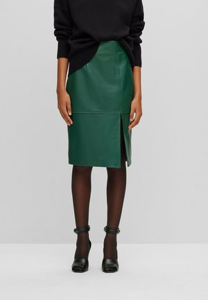 Femme portant une jupe en cuir vert foncé longueur genou avec une fente à l’avant, un pull noir à manches longues, des collants noirs et des escarpins noirs à bride à la cheville.