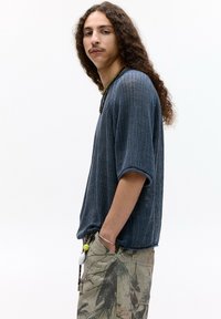 Cămașă tricotată cu dungi albastre, cu mânecă scurtă, purtată cu pantaloni cargo cu un model. Accesoriile includ un colier și un pandantiv decorativ pe pantaloni.