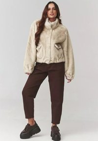 Kremowa kurtka z futerkiem syntetycznym z wysokim kołnierzem i elastycznymi mankietami, w zestawie z brązowymi spodniami woskowymi i czarnymi sneakersami na grubej podeszwie.