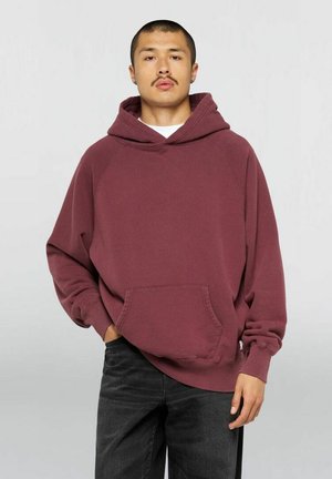 Giovane uomo con capelli corti e baffi che indossa una felpa bordeaux larga e pantaloni neri, in piedi con una mano in tasca.