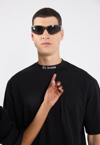 Sort oversized t-shirt med teksten "No drama" i hvid broderi på kraven, båret med futuristiske sorte solbriller.