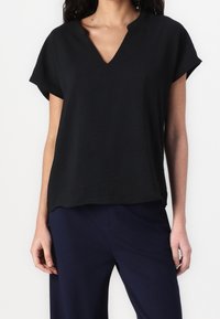 Top noir à manches courtes avec col en V, coupe ample, fabriqué en tissu léger. Texture subtile, ourlet arrondi, associé à un pantalon bleu marine.