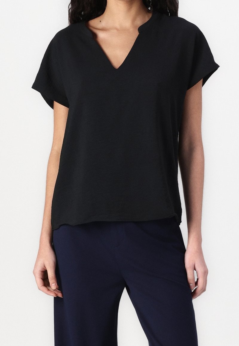 Top noir à manches courtes avec col en V, coupe ample, fabriqué en tissu léger. Texture subtile, ourlet arrondi, associé à un pantalon bleu marine.