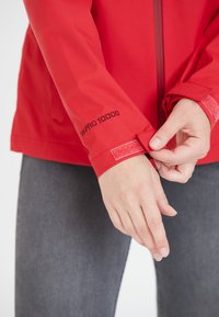 Veste imperméable rouge avec un col haut et des poignets ajustables. Présente le logo "WPRO 10000" sur la manche. Le tissu semble lisse et flexible.