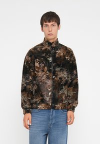Jachetă din fleece cu guler înalt, având un model de camuflaj multicolor în nuanțe de maro, verde și negru. Închidere cu fermoar, manșete elastice.