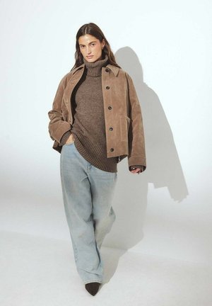 Braune Wildlederjacke über einem braunen Rollkragensweater, kombiniert mit hellblauen weiten Jeans und dunklen spitz zulaufenden Absätzen.