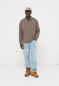 Brown long-sleeve polo shirt with a collar, light blue jeans, and tan work boots. The person is wearing a light-colored cap with text.