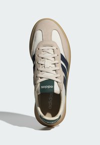 Béžová a bílá teniska s tmavě modrými pruhy, semišovým svrškem a zeleným logem na jazyku. Vybavena polstrovanou stélkou a gumovou podrážkou.