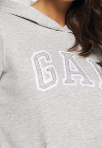 Sudadera gris con letras blancas en relieve que dicen "GAM" con una apariencia texturizada. El material tiene un aspecto suave y casual.
