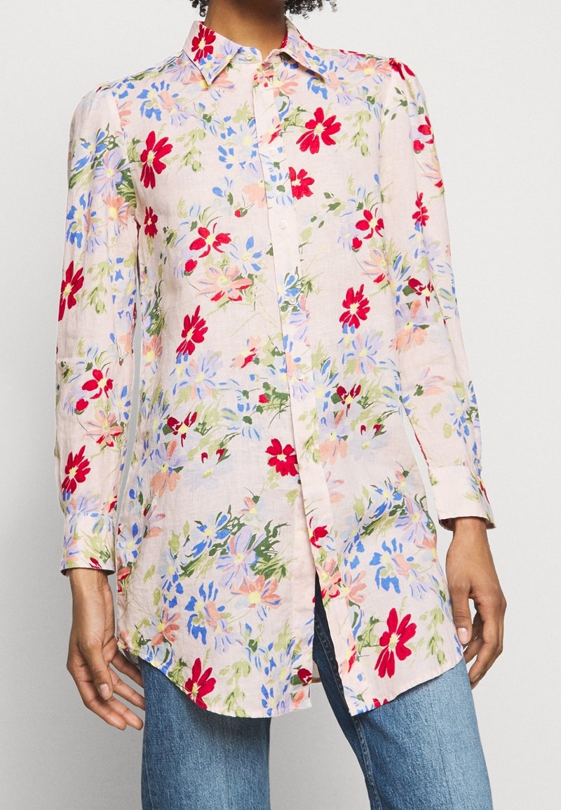 Chemise à manches longues, boutonnée, à motifs floraux rouges, bleus et roses, portée sur un jean bleu, visible du menton jusqu'à la mi-cuisse.