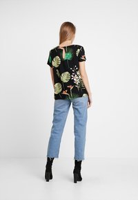 Top floreale nero con motivi di piante verdi e dettaglio a chiave sulla schiena, abbinato a jeans blu chiaro e stivali neri con tacco.