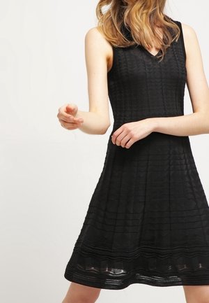 Femme portant une robe noire texturée sans manches avec un col en V, debout devant un fond clair uni, les bras en mouvement à mi-chemin.