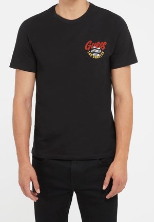 Mann trägt ein schwarzes Guess T-Shirt mit rotem und gelbem Logo-Schriftzug auf der linken Brust mit der Aufschrift "Guess Los Angeles Established 1981."