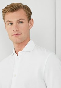Witte polo met een platte kraag, knoopsluiting en textiel met een textuur. Subtiel logo op de linkerborst, tegen een neutrale achtergrond.