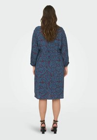 Sininen kuvioitu mekko, jossa on pitkät hihat ja vyötäröside, esittää punaista kukkakuviota. Yhdistetty mustiin korkokenkiin ja sileään materiaaliin.