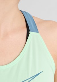 Miętowozielony sportowy top na ramiączkach z niebieskim paskiem z tkaniny. Posiada czarną wewnętrzną warstwę oraz geometryczny wzór w kolorze niebieskim na przodzie.