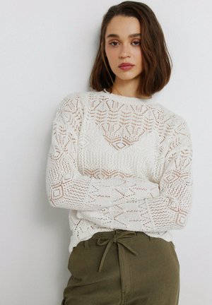 Woman with shoulder-length brown hair wearing a white patterned knit sweater and olive green pants with a tied waist.