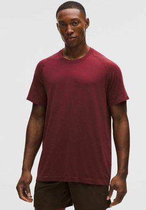 Ung mand stående iført en ensfarvet kortærmet vinrød skjorte og mørke shorts, neutral baggrund.