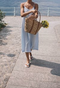 Kudottu olkihenkari ruskeilla nahkahandleilla, jonka henkilö pitää päällään vaaleansinisen mekon kanssa, kävellen kivipolulla veden lähellä.