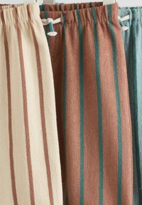Tři páry kalhot z měkké, texturované látky. Jeden pár je krémový s hnědými pruhy, další je rezavý s modrozelenými pruhy a poslední je světle modrý.