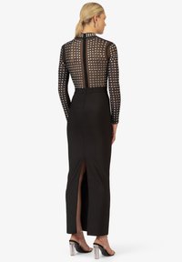Zwarte maxi-jurk met lange, doorschijnende mouwen. De achterkant heeft een ruitpatroon van zilveren studs. De rok heeft een split tot aan de dijen, aangevuld met doorzichtige hakken.