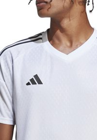 Weißes Sportshirt mit strukturiertem Muster, V-Ausschnitt und schwarzem adidas-Logo. Verfügt über schwarze Streifen auf den Schultern zur Akzentuierung.