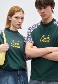 Δύο μοντέλα που φορούν σκουροπράσινα πουκάμισα «Carlo Colucci» με σχεδιασμένα μανίκια, το ένα κρατάει κίτρινη τσάντα, και τα δύο κοιτούν προς τα εμπρός.