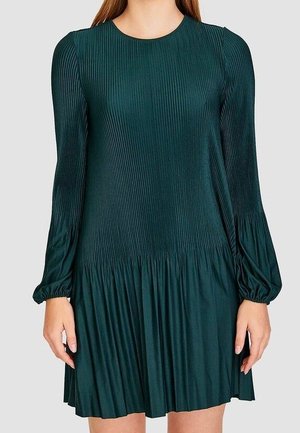 Woman wearing a dark green, long-sleeve pleated dress with a round neckline and gathered skirt hem.