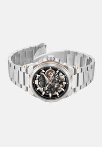 Srebrny zegarek ze stali nierdzewnej z czarną tarczą skeleton, różowozłotymi akcentami i bransoletą w formie ogniw. Posiada luminescencyjne wskaźniki godzinowe oraz koronkę.