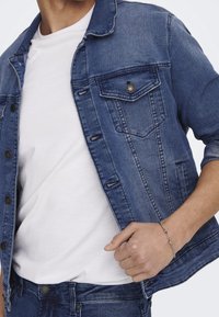 Spijkerjack in middenblauw met knoopsluiting, twee borstzakken en een casual pasvorm. Gedragen over een wit T-shirt, met een zichtbaar gestructureerde stof.