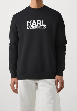 Sort langærmet sweatshirt med hvid "KARL LAGERFELD" logo og silhouet grafik, båret med hvide bukser på en stående person.