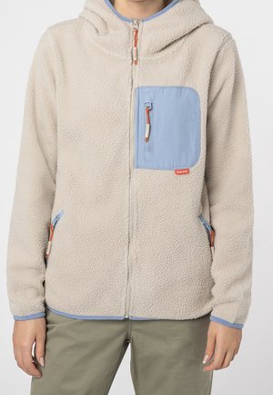 Veste polaire beige à fermeture éclair avec une poche poitrine bleue et des poignets assortis bordés de bleu, portée avec un pantalon vert olive.