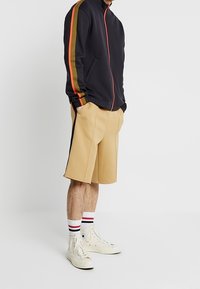 Persona que lleva una chaqueta negra con cremallera y franjas naranjas en las mangas, pantalones cortos beige hasta la rodilla, zapatillas blancas de caña alta y calcetines rayados.