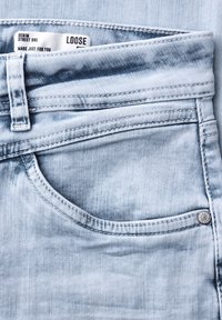 Jasnoniebieska kieszeń dżinsów z widoczną pętlą na pasek, przeszyciami, metalowym nitami oraz częściowo widoczną białą metką z napisem "Loose Fit".