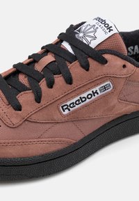 Reebok tenisice s gornjim dijelom od smeđeg velura, crnim vezicama i gumenom potplatom. Imaju oznaku s logom i teksturirani dizajn sa strane.